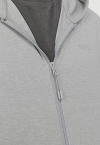 Sudadera con cremallera de color gris claro hecha de material suave, que presenta una cremallera lisa y un logo tonal cerca del cuello, llevada sobre una camiseta oscura.