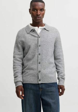 Homme portant un cardigan gris à boutons sur une chemise blanche et un jean bleu, debout devant un fond clair uni.