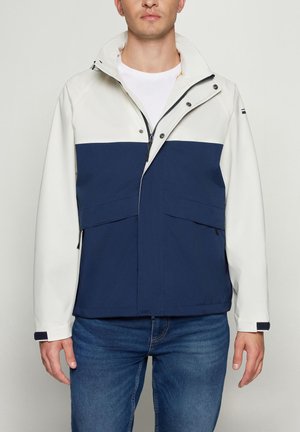 Mand iført en hvid og mørkeblå lynlåstrøje med to store forlommer, kombineret med blå jeans på en ensfarvet baggrund.