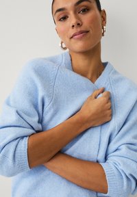 Pull en laine bleu clair avec un col en V, des poignets côtelés et une texture douce. Le modèle tient le pull près du corps.