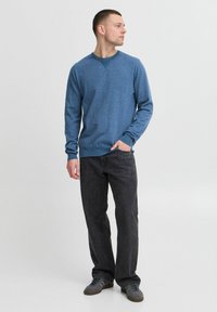 Blauwe gebreide sweater met lange mouwen en V-hals, gecombineerd met donkere spijkerbroek en grijze sneakers. Zachte textuur en relaxte pasvorm.