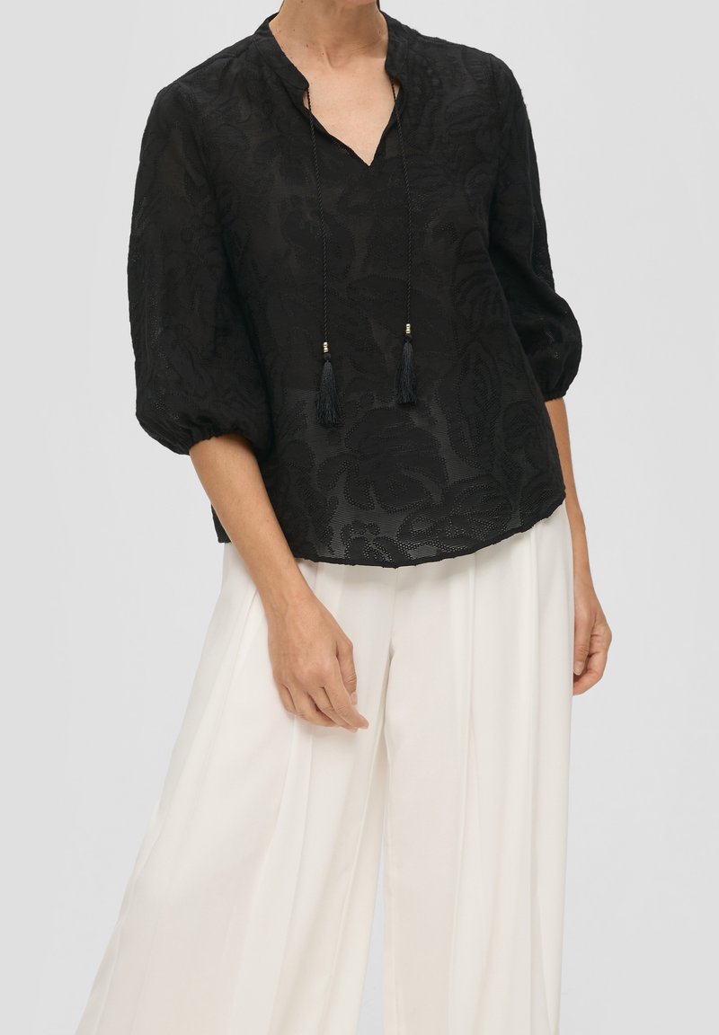 Blusa negra con estampado floral, mangas 3/4, escote en V y lazos de borlas. Combinada con pantalones anchos blancos. Tejido ligero y texturizado.