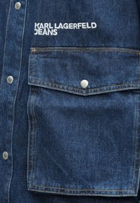 Zbliżenie na kieszeń niebieskiej kurtki dżinsowej z metalowym zatrzaskiem i białym haftowanym logo "Karl Lagerfeld Jeans" nad kieszenią.