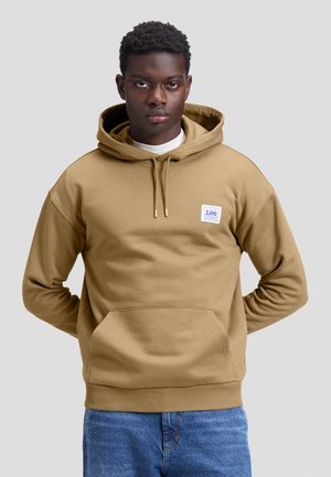 Joven con sudadera con capucha color tostado con bolsillo delantero y pequeño logo de Lee en el pecho, de pie con las manos detrás de la espalda.
