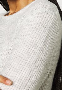 Ljusgrå stickad tröja med rund hals och texturerat mönster, med en mjuk, lös väv och synliga sömnadsdetaljer.