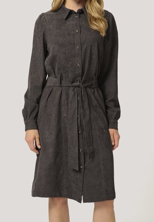 Femme portant une robe gris foncé à manches longues, boutonnée, avec un col et une ceinture en tissu nouée à la taille, debout devant un fond uni.