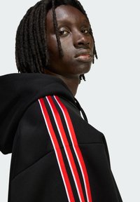 Čierna mikina s kapucňou s červenými a bielymi pruhovanými detailmi na rukávoch. Má voľný strih a textúrovanú látku. Bez ďalších ozdôb.