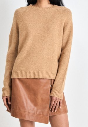 Vrouw draagt een zacht, kabelgebreid trui in camelkleur, gecombineerd met een bruine leren asymmetrische wikkelrok met een ritssluiting aan de zijkant.