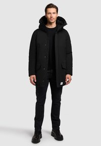 Czarna pikowana kurtka z kapturem, zapinana na suwak i napy, z dwoma przednimi kieszeniami oraz tkawaną metką przy dolnym brzegu. Noszona na czarnym swetrze.