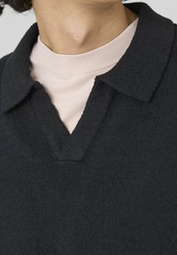 Gros plan sur une personne portant un t-shirt crewneck rose clair sous un pull en tricot noir avec un col en V.