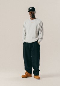 Mees, kellel on seljas heledashall dressipluus, mustad püksid, beežid saapad ja must New York Yankeesi pesapallikork, seisab käed taskutes.