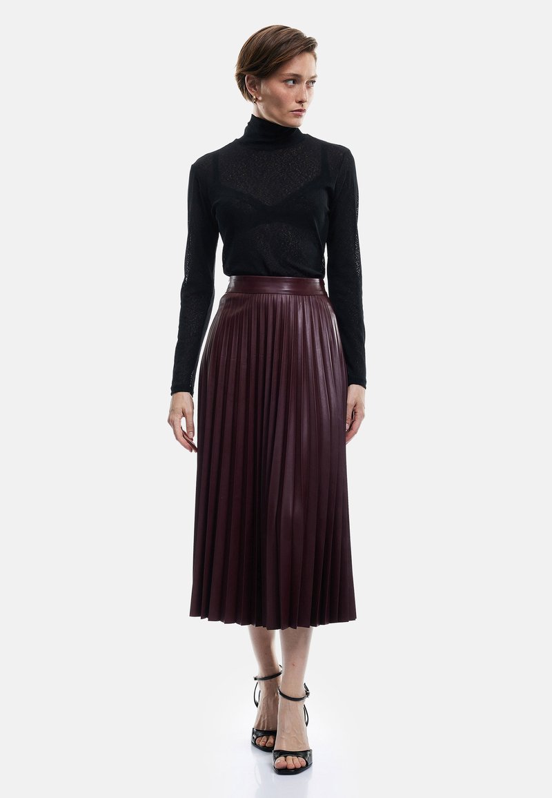 Haut long à manches noires avec un motif texturé, associé à une jupe midi plissée en simili cuir bordeaux et à des escarpins noirs à brides.
