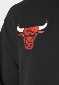Sweatshirt noir avec un logo de taureau rouge et blanc brodé sur la poitrine gauche, avec des détails distincts sur le visage de l'animal.