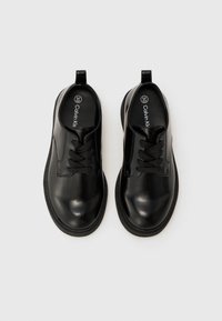 Chaussures à lacets en cuir noir avec une finition brillante, présentant un bout rond, des lacets noirs et un passant de talon en boucle. Texture lisse avec une semelle contrastée.