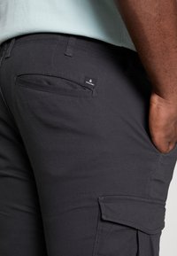 Black cargo pants with a side pocket and a small brand tag. Smooth fabric texture and fitted design. Shown next to a hand in pocket.