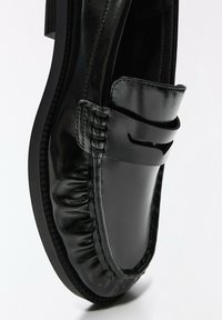 Chaussure loafer en cuir noir brillant avec des détails cousus et un design moc toe sur un fond blanc uni.
