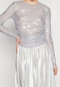Durchsichtige silberne Bluse mit langen Ärmeln und funkelnder Textur, gerafftem Bund, kombiniert mit einem plissierten silbernen Rock, der den glänzenden Stoff und das sanfte Design hervorhebt.
