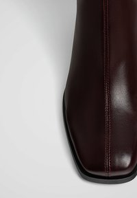 Burgundy nahkasaappaat, joissa on kiiltävä pinta, neliönmuotoinen varvas ja kontrastivärinen ommel sivussa. Musta teksturoitu pohja.