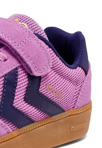 Różowy zamszowy sneaker z granatowymi akcentami, paskiem na rzep, fakturowaną powierzchnią i brązową gumową podeszwą. Posiada zygzakowaty wzór i złote detale z logo.