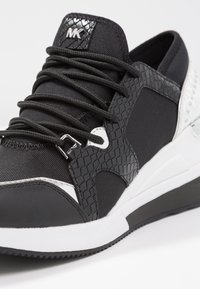 Svart sneakers med en strukturerad ovandel, kontrasterande vita detaljer och ett mönstrat sidopanel. Har snören och en logotyp på tungan.