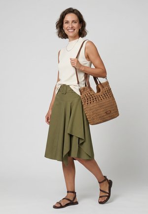 Lachende vrouw die een mouwloze witte top draagt, een asymmetrische olijfgroene rok, bruine sandalen met bandjes en een gevlochten beige handtas over haar schouder draagt.