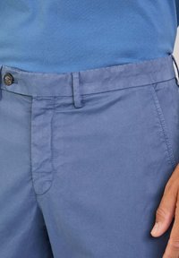 Pantalones de tejido ligero en azul claro, con una textura suave, que cuentan con una cinturilla estándar, bolsillos frontales y un cierre de botón.