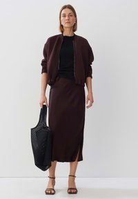 Dunkelburgunderfarbener Bomberjacke, schwarzes Oberteil, passender Rock mit seitlichen Schlitzen, schwarze Netztragetasche und braune Sandalen mit Absatz. Glatte Texturen überall.