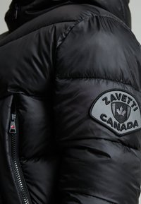 Veste noire en duvet avec des panneaux matelassés, une poche zippée et un écusson circulaire portant "ZAVETTI CANADA" et un motif de feuille.