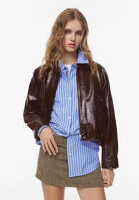 Veste en cuir marron à finition brillante, portée sur une chemise rayée bleu et blanc. Le mannequin porte également une jupe marron texturée.