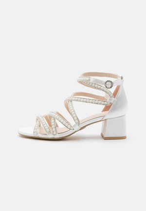 White satin heeled sandals featuring multiple crisscross straps embellished with pearls. Block heel design with a rounded toe.