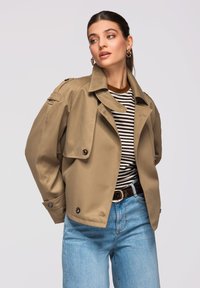 Femme portant un trench-coat beige par-dessus une chemise rayée noir et blanc et un jean bleu clair, regardant sur le côté devant un fond uni.