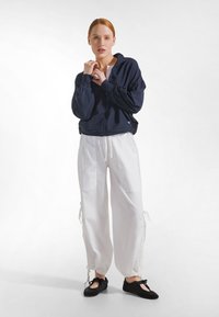 Marineblå lynlåsjakke med løs pasform, kombineret med hvide løstsiddende bukser med sidelommer og snørelukning ved benene. Sorte flade sko.