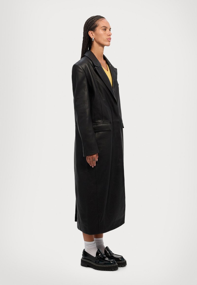 Black leather long coat with a notched collar, side pockets, and a textured finish. Worn over a yellow top with dark shoes and white socks.