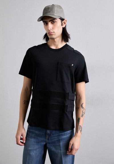 T-shirt nera con taschino sul petto e tre strisce orizzontali, tessuto texturizzato, abbinata a jeans di denim blu. Il modello indossa un cappellino grigio chiaro.