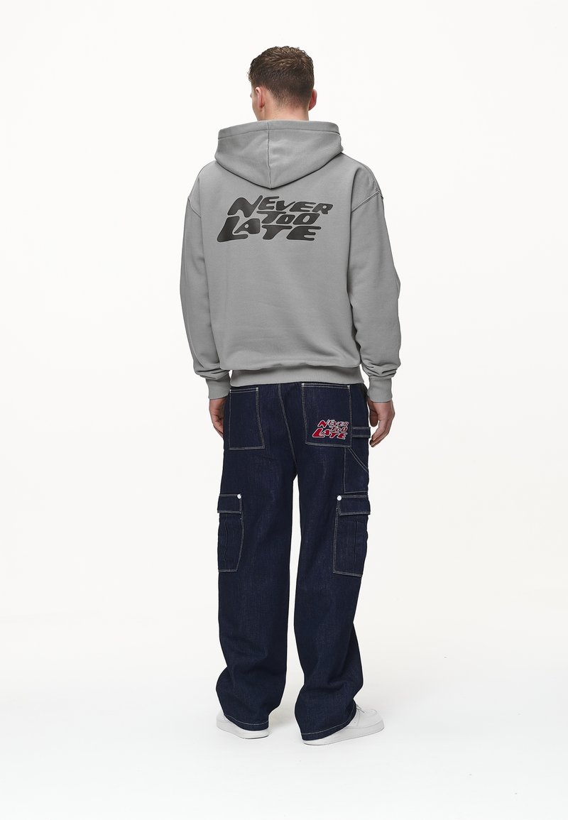 Szara bluza z kapturem z nadrukiem "NEVER TOO LATE" na plecach. Do tego ciemne dżinsowe spodnie cargo z bocznymi kieszeniami i detalem z logo.