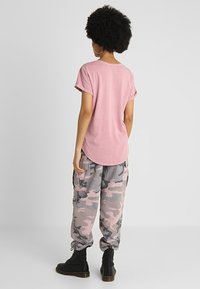 T-shirt rosa a maniche corte con orlo curvo, abbinato a pantaloni cargo grigi con camouflage. L'outfit presenta una vestibilità rilassata e dettagli minimi. Stivali neri completano il look.