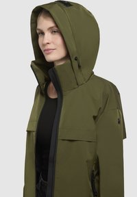 Veste imperméable vert olive avec col haut, capuche réglable et fermeture éclair noire. Dispose de poches avant et d'une texture lisse et mate.