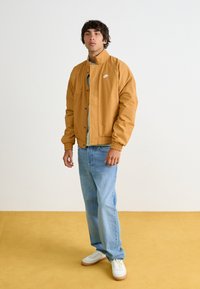 Giacca bomber marrone chiaro con polsini elasticizzati e un piccolo logo sul petto, abbinata a jeans azzurri e sneakers bianche.
