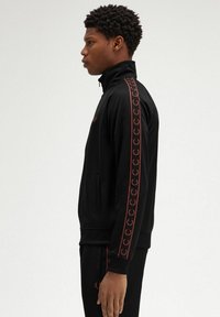 Giacca nera con zip realizzata in tessuto liscio, con colletto alto, tasche laterali e loghi rossi lungo le maniche e il petto.