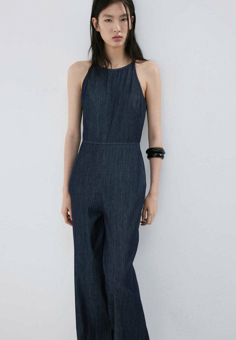 Femme aux longs cheveux foncés portant une combinaison sans manches en jean foncé et un bracelet noir, debout devant un fond clair uni.