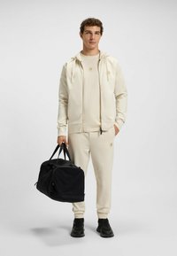 Beige hoodie och matchande joggers tillverkade av en slät tyg, tillsammans med en svart duffelväska. Outfiten har en logotyp på bröstet och byxorna.