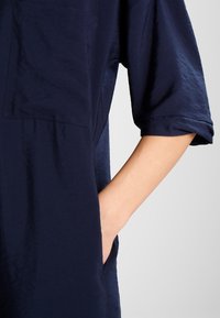 Indumento blu navy con una vestibilità ampia, maniche corte, una tasca quadrata sul petto e tasche laterali. Tessuto liscio e leggermente strutturato.