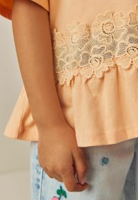 Pfirsichfarbene Baumwollbluse mit Rüsche am Saum und cremefarbenen Spitzenblumenapplikationen. Die Hand liegt auf hellblauen bestickten Jeans.