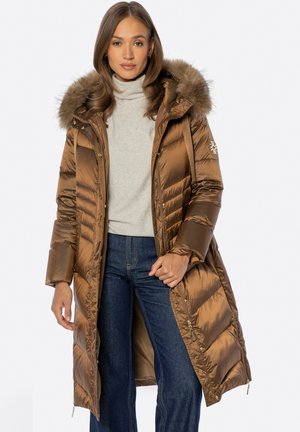 Frau trägt eine lange braune Steppjacke mit Pelzkapuze über einem hellgrauen Rollkragenpullover und blauen Jeans, steht vor einem schlichten Hintergrund.