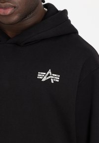 Schwarzer Hoodie mit weicher Textur, der ein auffälliges silbernes Grafiklogo auf der Brust trägt, das geometrische Linien und Formen zeigt.