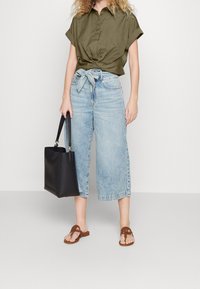 Camicia oliva a maniche corte con chiusura a bottone, jeans a gamba larga blu chiaro, sandali piatti marroni e una borsa tote nera.