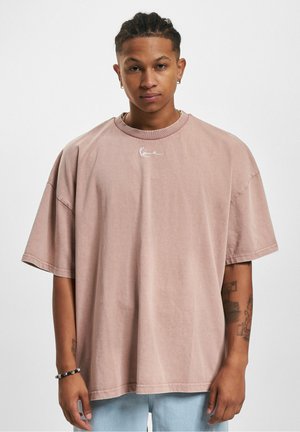 Oversized light pink cotton t-shirt with short sleeves, round neck, and subtle white logo detailing on the chest. Smooth texture.