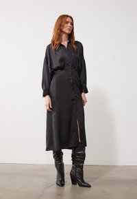 Sort satin kjole med lange ærmer, knaplukning fortil og sideslids. Parret med knæhøje sorte læderstøvler. Glat, skinnende tekstur.
