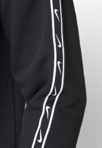 Felpa nera con strisce laterali bianche e loghi Nike ripetuti. Tessuto morbido, design aderente che enfatizza la forma delle braccia.