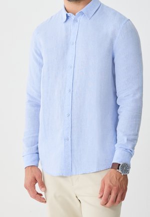 Mand iført en lyseblå, langærmet skjorte med knapper og beige bukser med et armbåndsur på venstre håndled, stående mod en hvid baggrund.